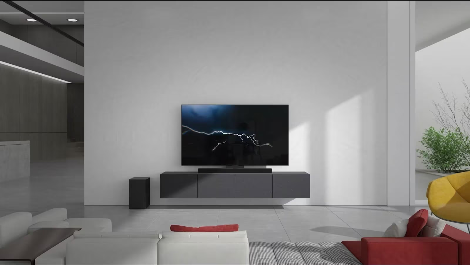 The Soundbar  is placed on gray cabinet with a TV in the living room. A black wireless subwoofer is placed on the floor on the left side and the sunlight comes in from the right side of the picture. A white and red colored long sofa is placed facing the TV and Soundbar .